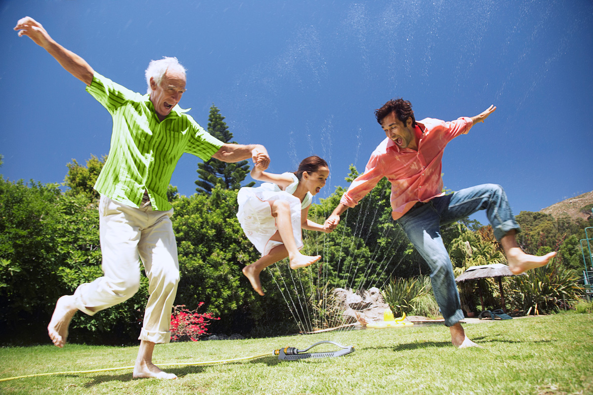 Grandfather, Father, and Daughter jumping through water sprinkler.